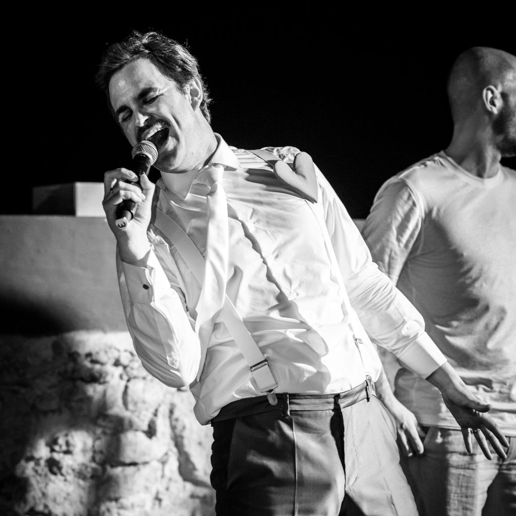 BingoShow Bingo Show de Fede Arias. Hombre cantando con micrófono, vestido con camisa blanca y tirantes, en un escenario. Fondo oscuro con otra persona al lado.
