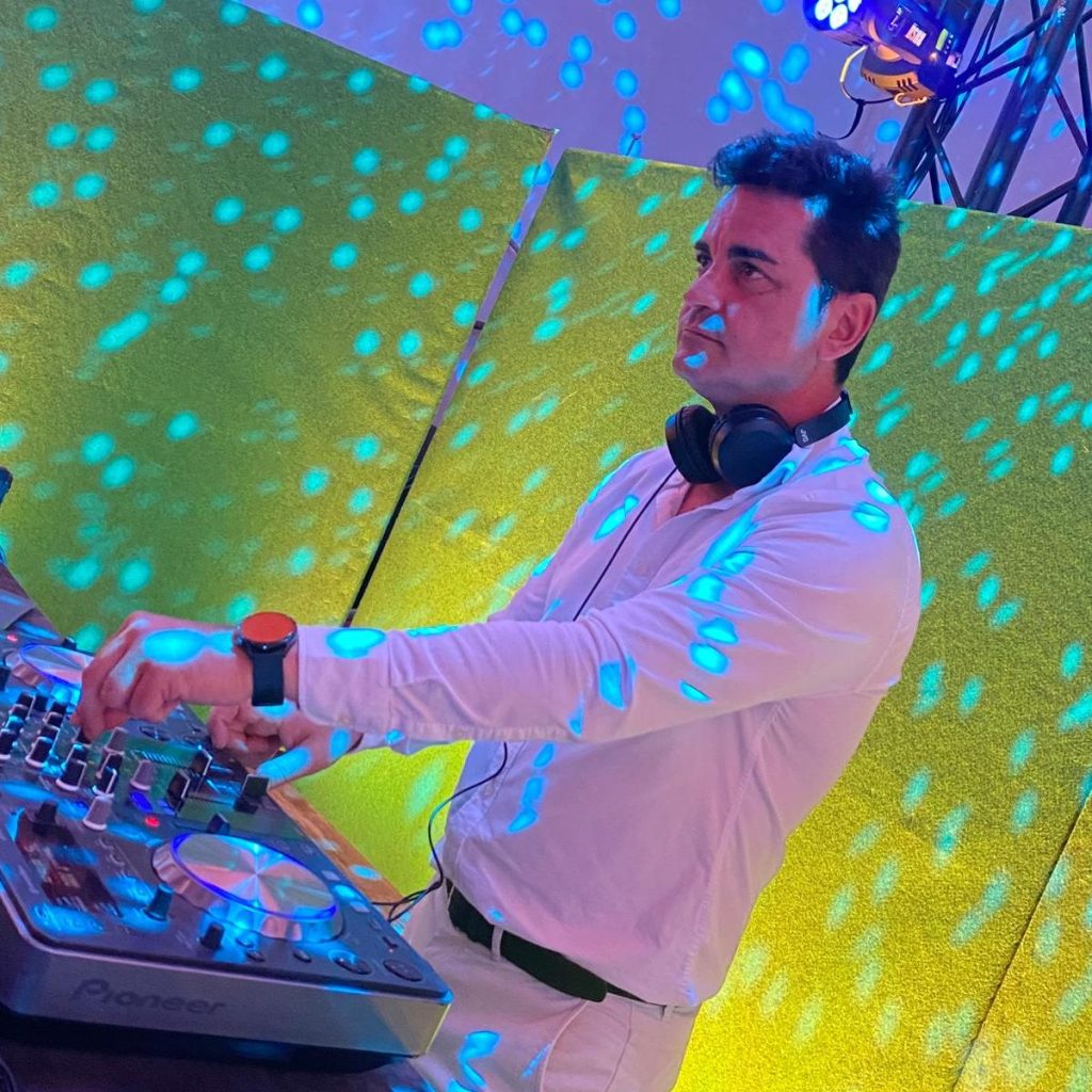 BingoShow Bingo Show de Fede Arias. Un DJ tocando en un evento, con luces de colores brillantes proyectadas en la pared y en su ropa. Luce concentrado mientras manipula el equipo de mezcla en su mesa.